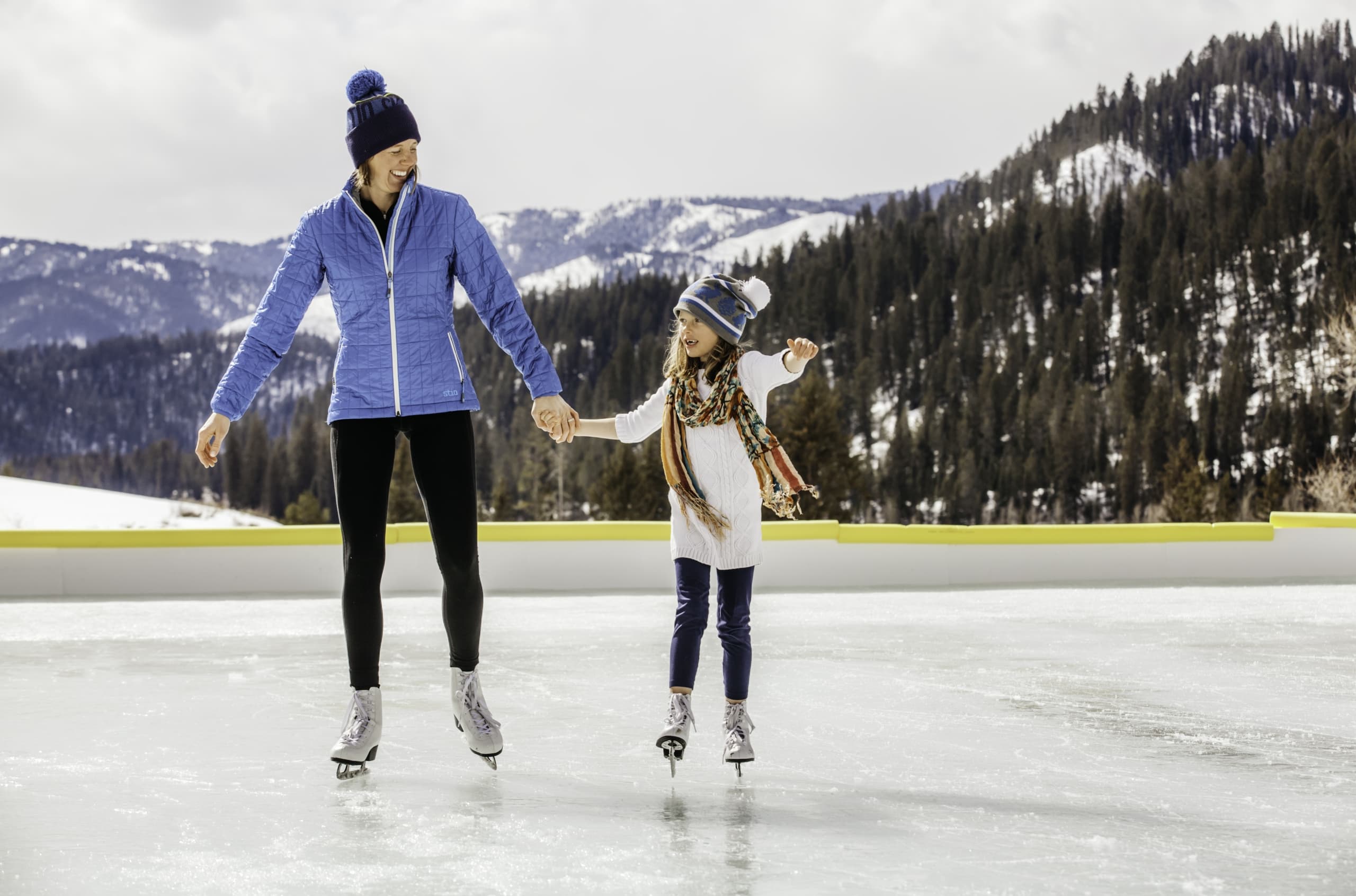 A mother and child ice skating outdoors surrounded by mountains